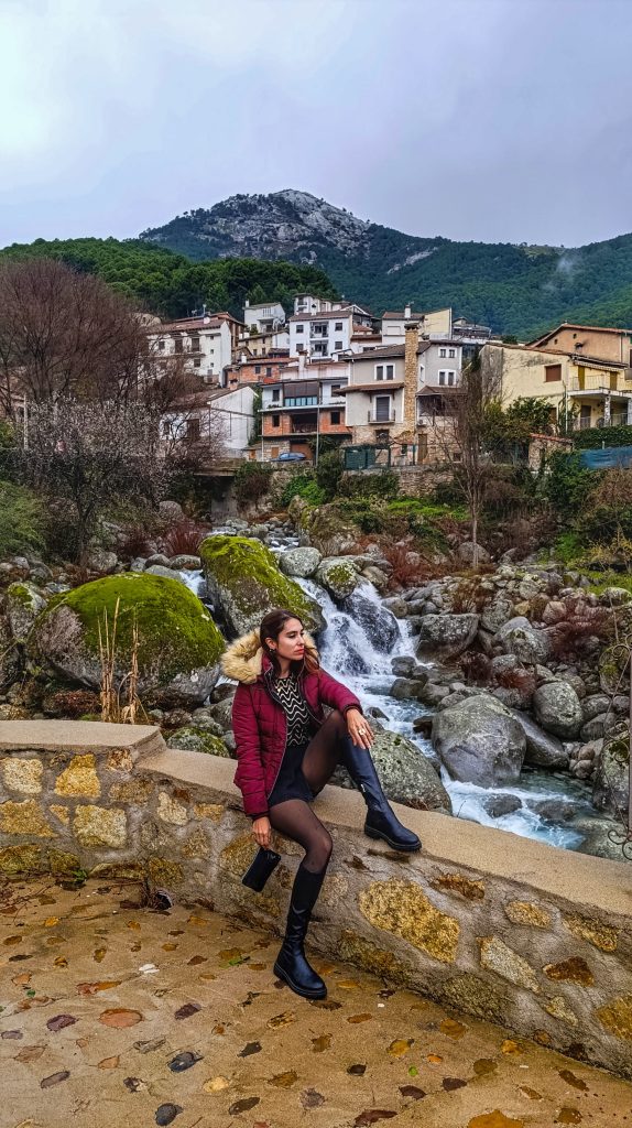 Vista del pueblo de Guisando en la Sierra de Gredos, con un salto de agua en primer plano y las casas de piedra al fondo, envueltas en un cielo nublado que resalta el paisaje rural