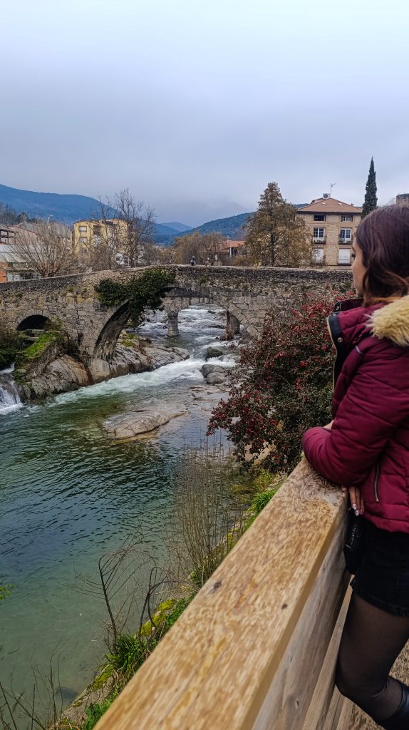 Vista de Arenas de San Pedro con su puente medieval de piedra en primer plano, el río fluyendo bajo sus arcos y las montañas de Gredos al fondo en un día nublado