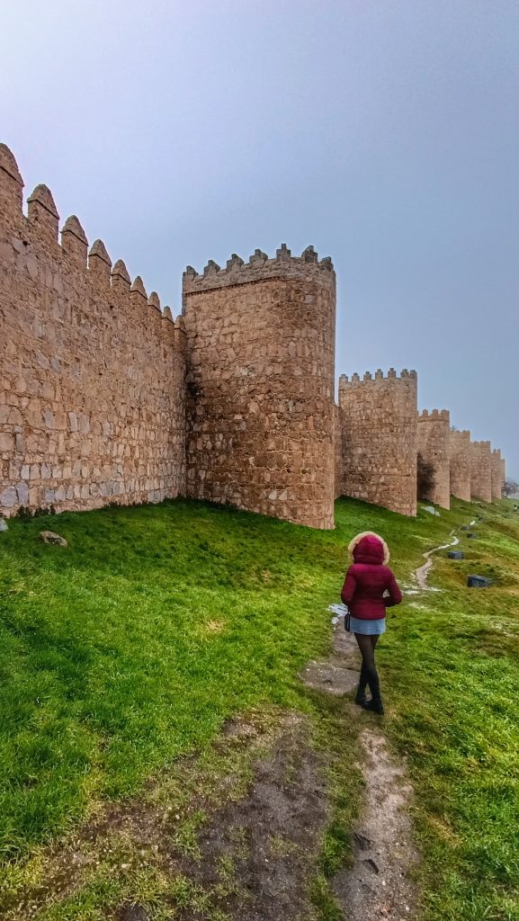 Imponentes murallas de Ávila bajo un cielo nublado, con una chica con capucha admirando la histórica fortificación.