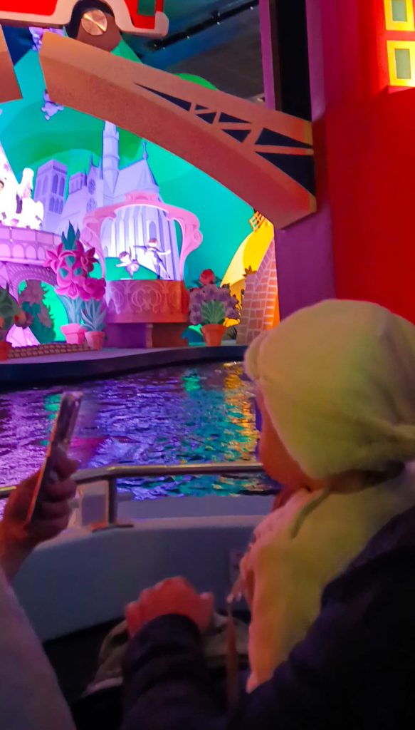 Bebé sentado en la atracción "It's a Small World" de Disneyland París, rodeado de coloridas decoraciones y figuras representando distintas culturas del mundo.